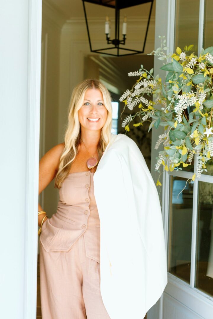 Smiling woman in a beige dress with a white jacket over her shoulder.