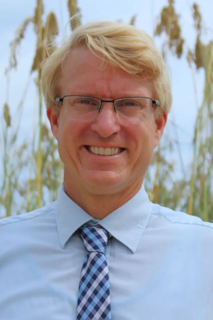 Smiling man with glasses in a blue shirt and striped tie outdoors.