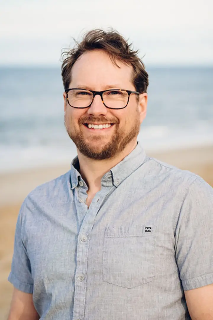 Smiling man with glasses standing on a beach.