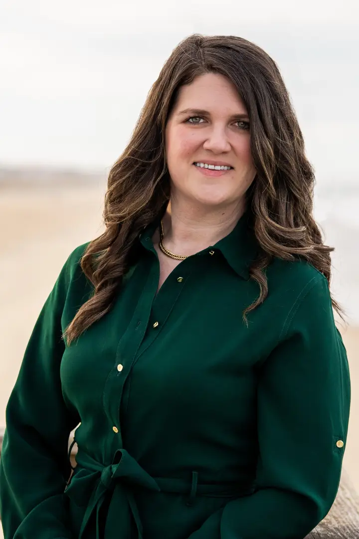 Woman with long wavy hair wearing a dark green blouse smiles outdoors.